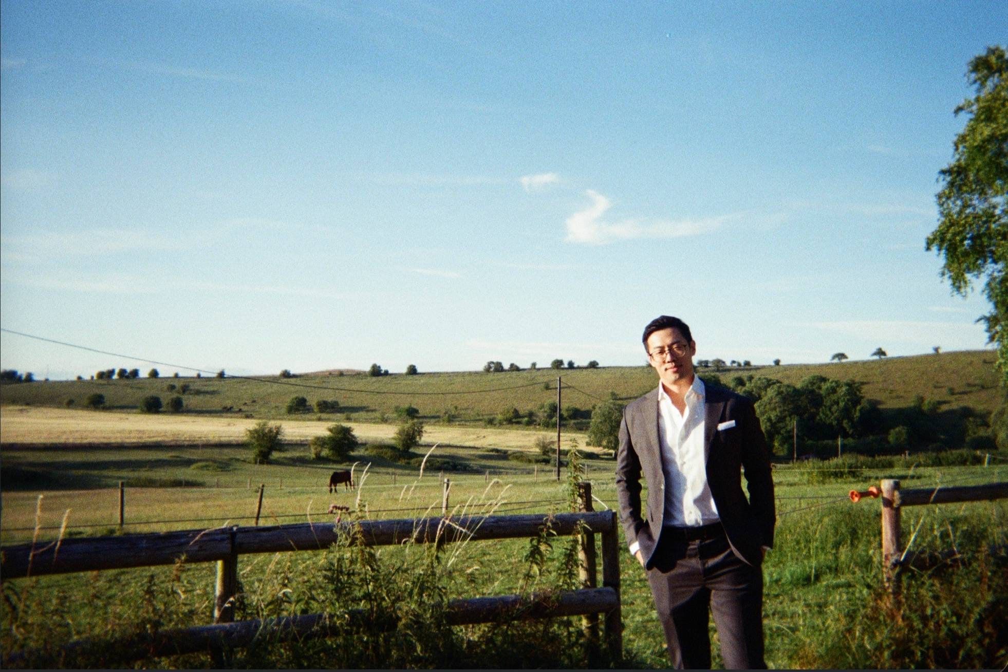 Jerry Zheng standing beside a wooden fence, with open farmland and a grazing horse behind him.